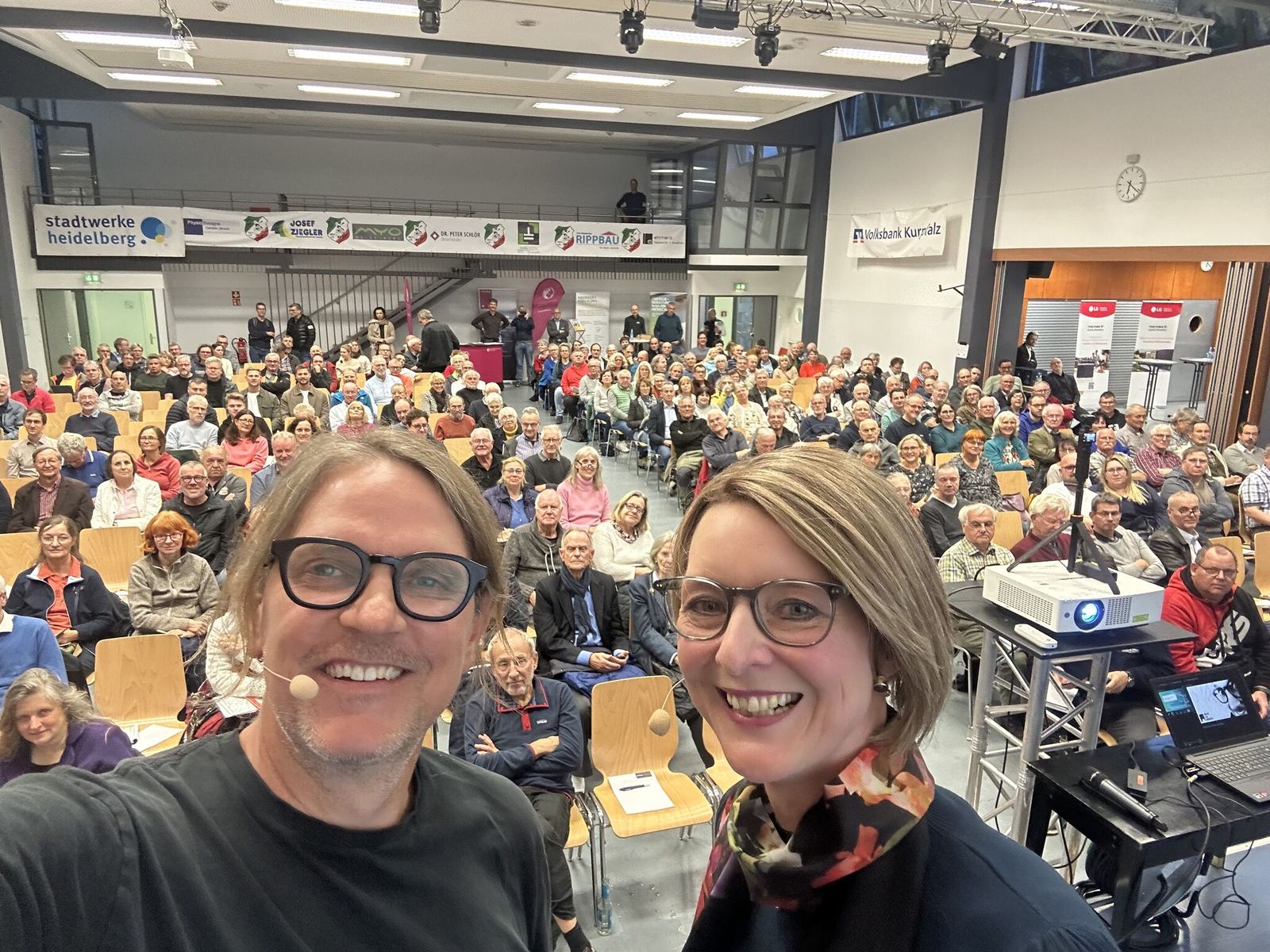 Carsten Herbert und Anja Floetenmeyer-Woltmann machen ein Selfie mit dem vollen Saal beim Wärmepumpen-Infotag in Heidelberg