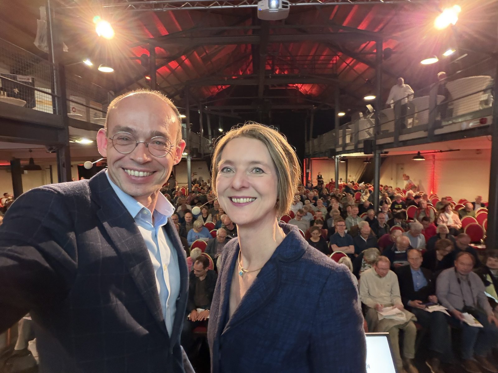 Dr. Marek Miara und Anja Floetenmeyer-Woltmann auf der Bühne der Jabelmannhalle, hinter ihnen das voll besetzte Publikum
