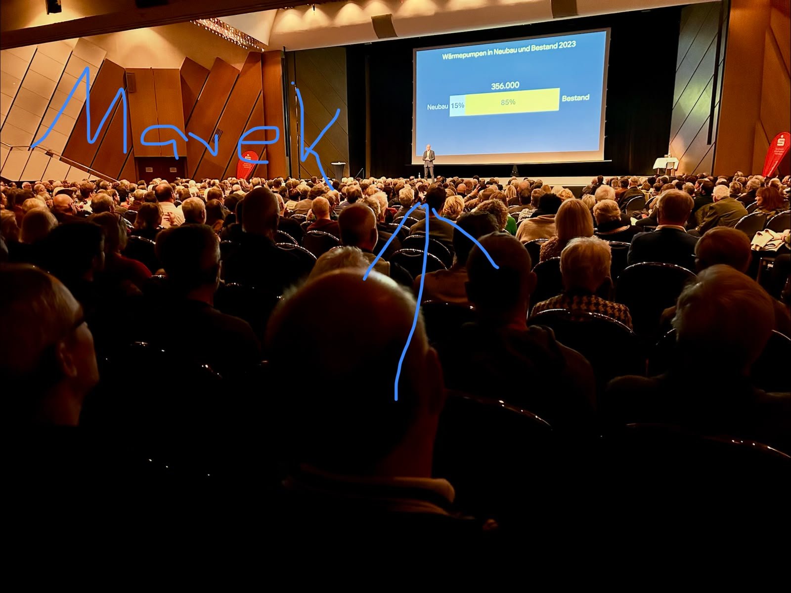 Blick aus dem vollen Saal auf die Bühne, Dr. Marek Miara mit Pfeil und Namen markiert